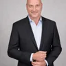 Peter DeCaprio in a dark jacket and open collar shirt, adjusting his cuff for a studio portrait