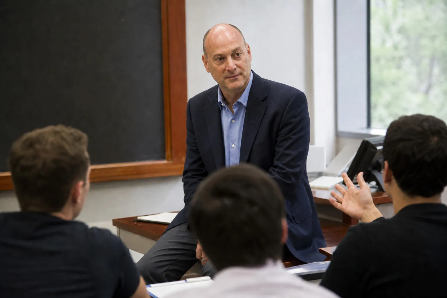 Peter DeCaprio seated on the edge of a desk at the front of a classroom, listening to a student's question with a blackboard behind him